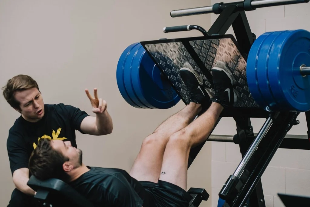 Coach in MDK-shirt begeleidt een klant op de leg press