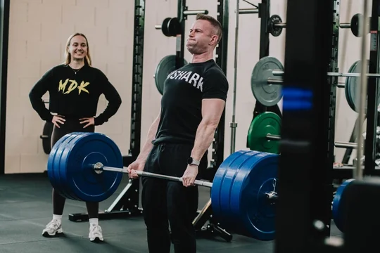 Coach in MDK-trui begeleidt een klant bij een deadlift