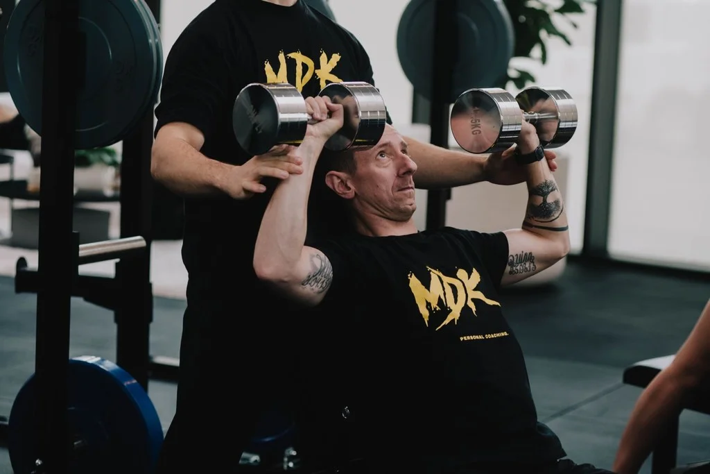 Coach ondersteunt een klant bij een schouderpress met dumbbells
