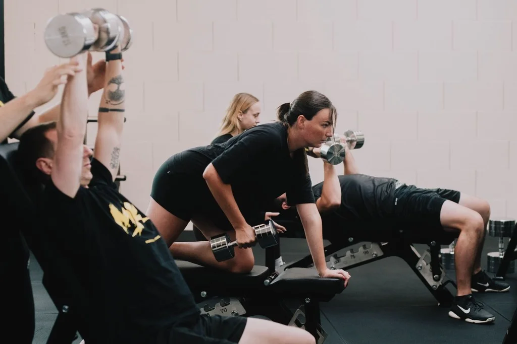 Personal training bij MDK: twee klanten trainen met dumbbells op een bank, met hun coach erbij